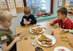 Kilkoro dzieci przy stole ozdabia pierniki przy wykorzystaniu lukru oraz posypki.